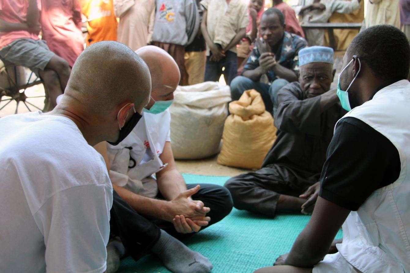 A group of men sit in a circle outside, speaking to each other. 