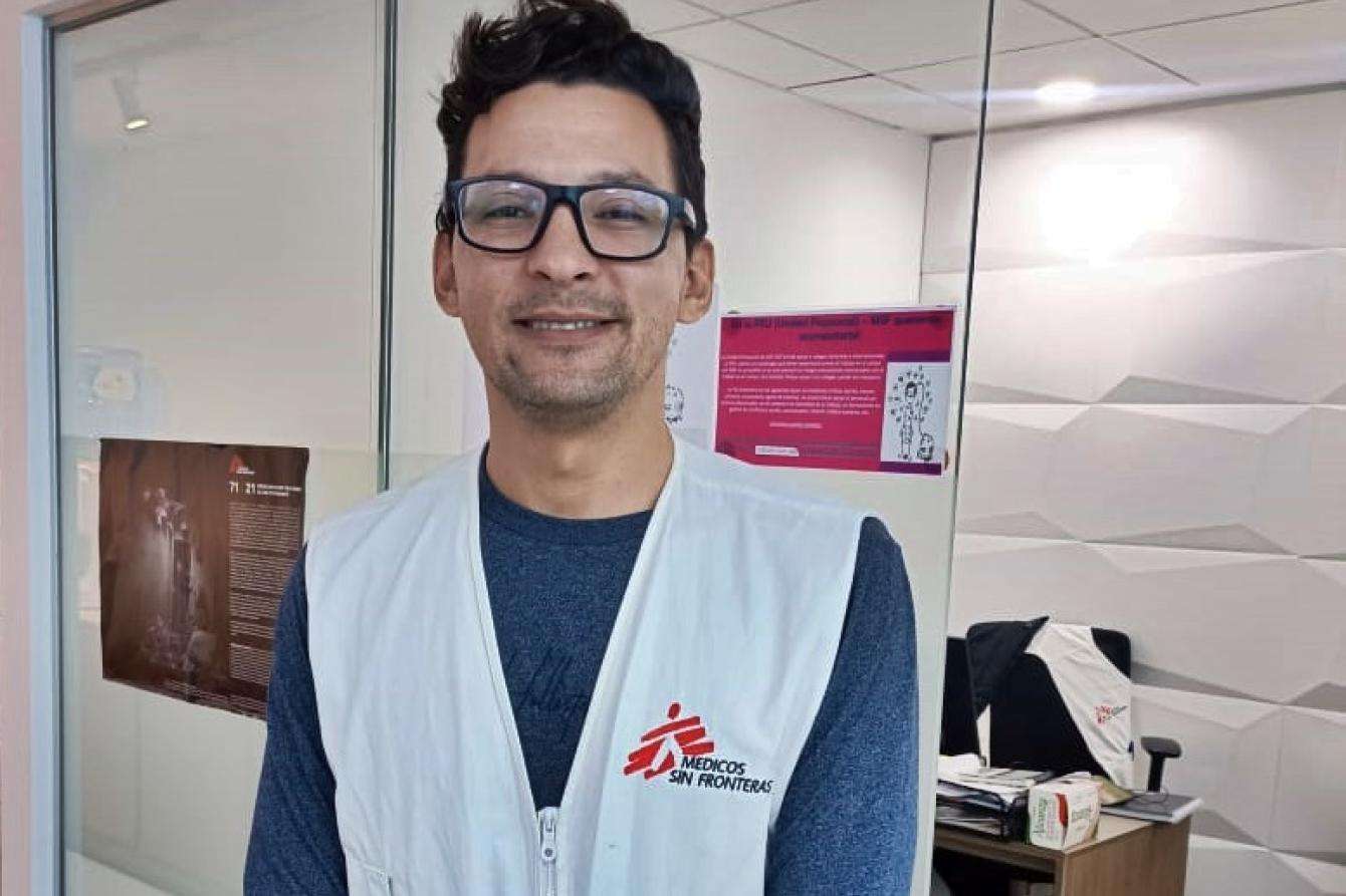 Portrait of a man with brown hair and black glasses, wearing an MSF vest in an office setting and smiling.