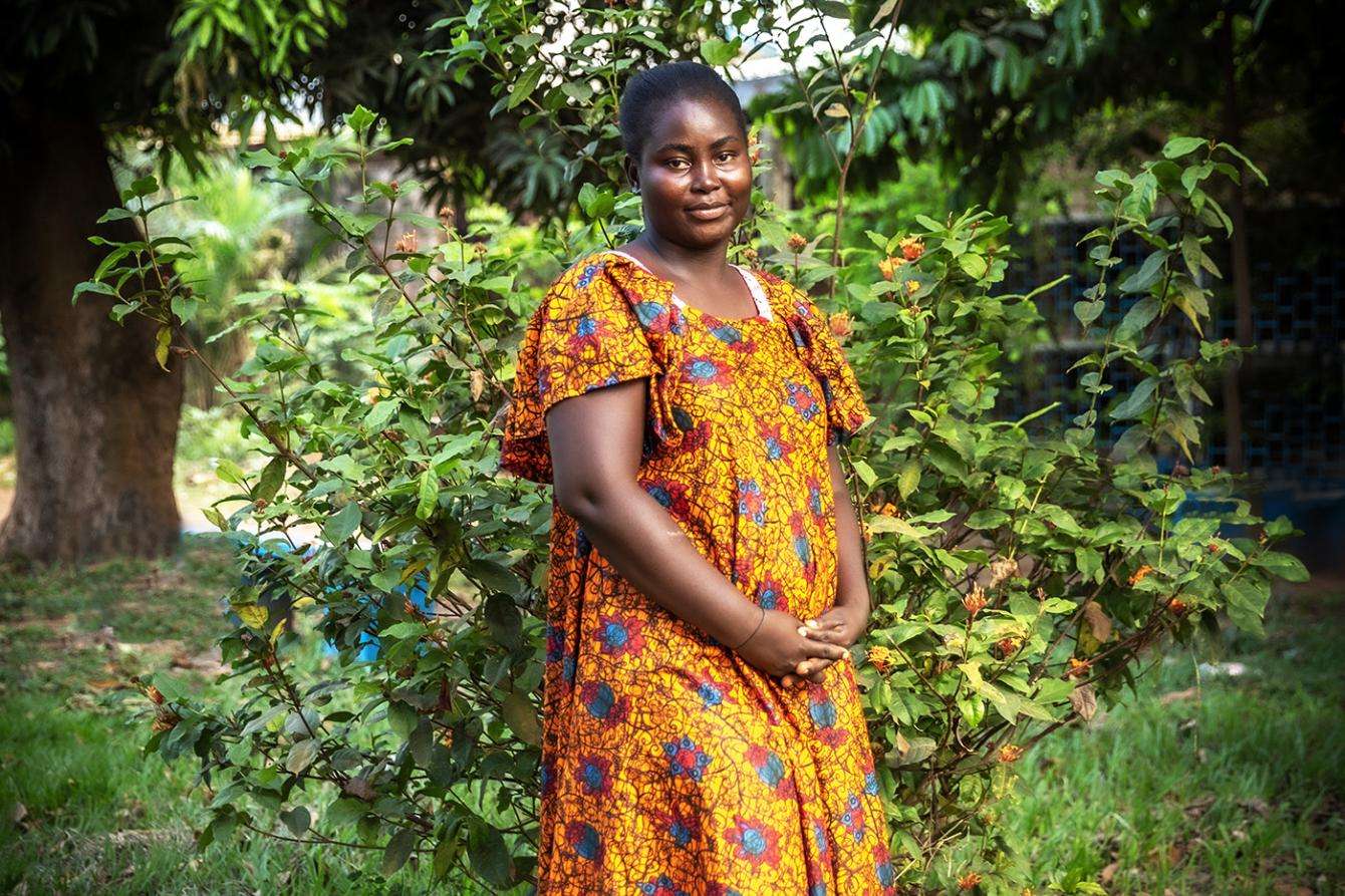 A young woman in an orange colorful dress stands in a green area outside in the Central African Republic. 