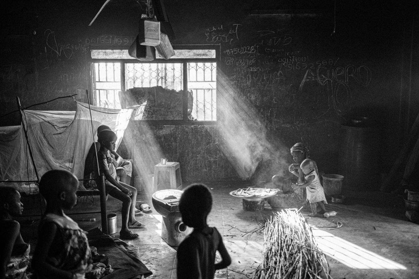 The inside of an abandoned house, adults and children sit, and light streams in through the window.